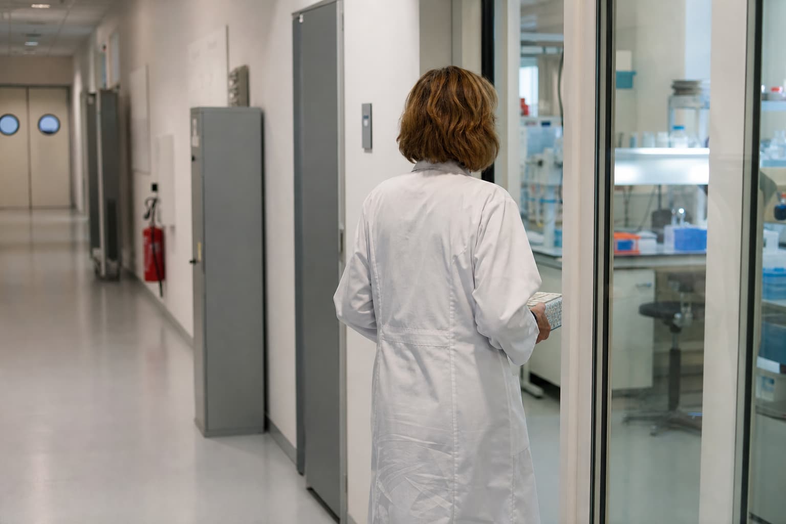 Illustration générée par IA : corridor sobre d’un centre de recherche, avec une technicienne de dos transportant une boîte d’échantillons vers une salle vitrée.