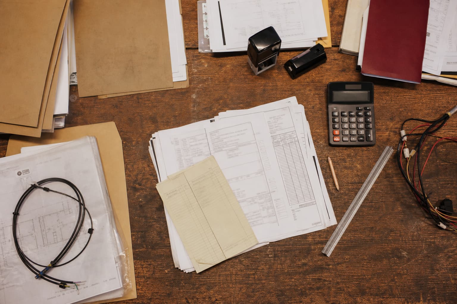 Illustration générée par IA : vue en plongée d’une table d’atelier usée avec bon de commande, calculatrice, câbles, tampon dateur et chemises d’approbation.
