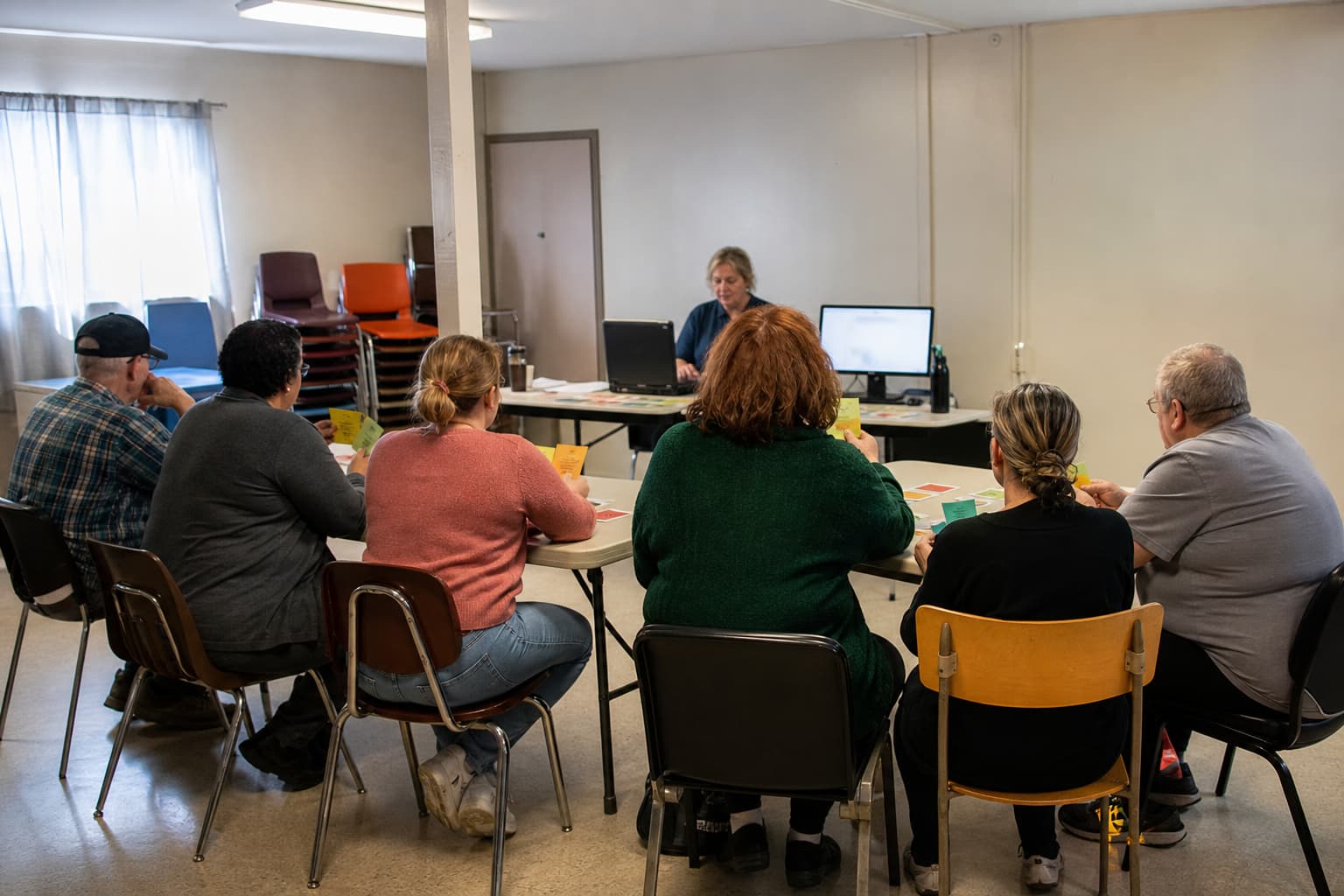 Illustration générée par IA : groupe d’adultes de différents âges vu de dos dans un local associatif, pendant une formation sur les accès numériques.