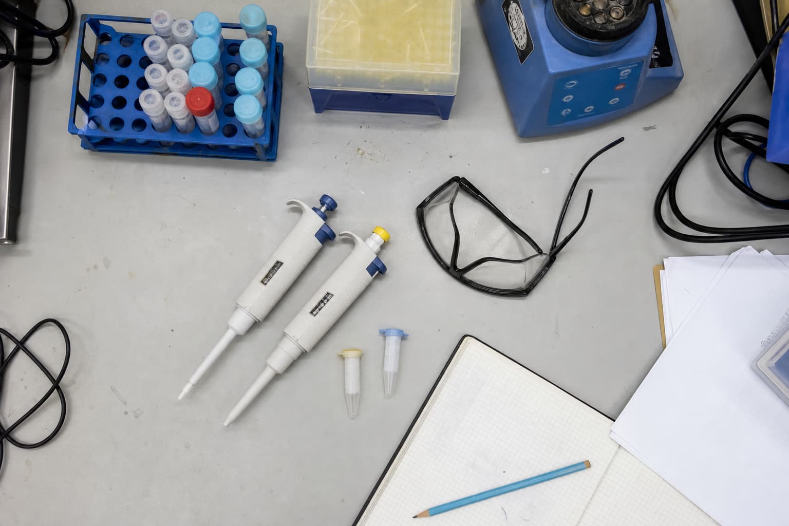 Illustration générée par IA : vue en plongée d’une table de laboratoire utilisée, avec carnet fermé, pipettes, câbles, lunettes et échantillons.